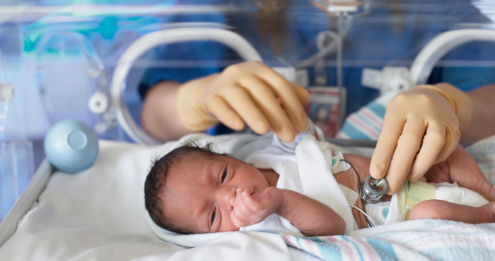 Image is of a baby in a NICU incubator, laying on a hospital blanket. A nurse's gloved hands are in the incubator, holding a stethoscope to the baby's exposed belly.