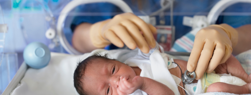 Image is of a baby in a NICU incubator, laying on a hospital blanket. A nurse's gloved hands are in the incubator, holding a stethoscope to the baby's exposed belly.